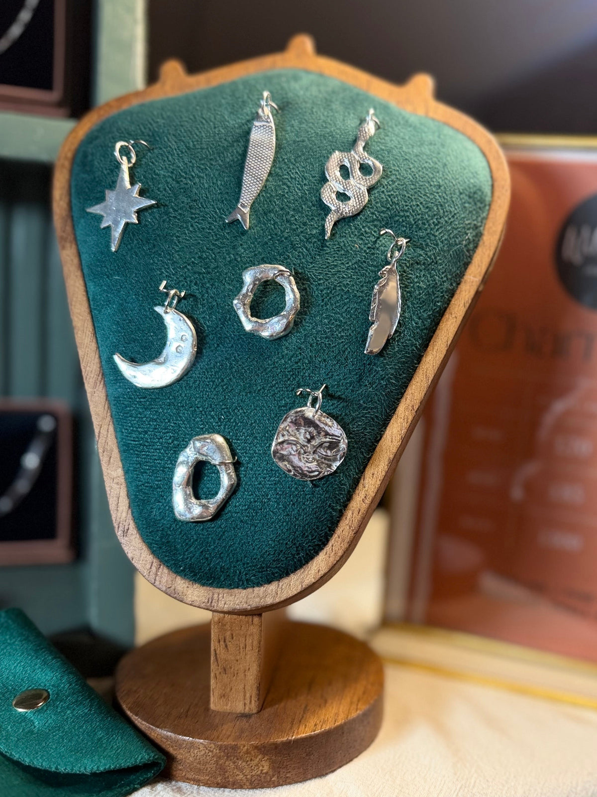 Handmade maxi silver charms displayed on a dark green velvet and wooden jewellery bust, featuring a snake, crescent moon, star, fish, floral designs and organic textured talismans by Wild One Jewellery.