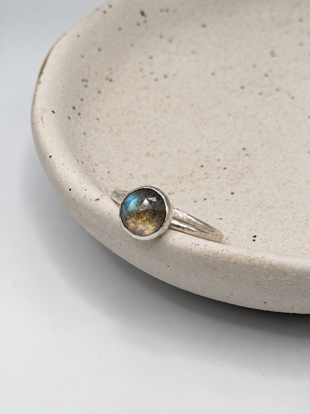 Handcrafted recycled sterling silver labradorite ring with a round faceted gemstone showing blue and golden flashes, displayed on a speckled ceramic dish.