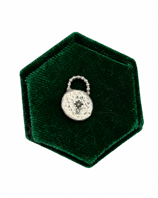 Handmade recycled sterling silver Love Lock charm featuring a handstamped pole star in the centre of a textured, chunky silver disc, with a beaded detail across the top, displayed on a deep green velvet jewellery cushion.
