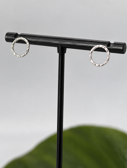 Pair of handmade recycled 925 sterling silver circle stud earrings with a delicate dimple texture, displayed on a black jewellery stand against a soft neutral background with a green leaf.